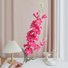 Une main tient une tige de fleurs artificielles de delphinium roses contre un fond de rideau rose pâle.