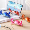 A pink crab-shaped desk organizer with a small phone holder is shown next to a white box and eyeglasses.