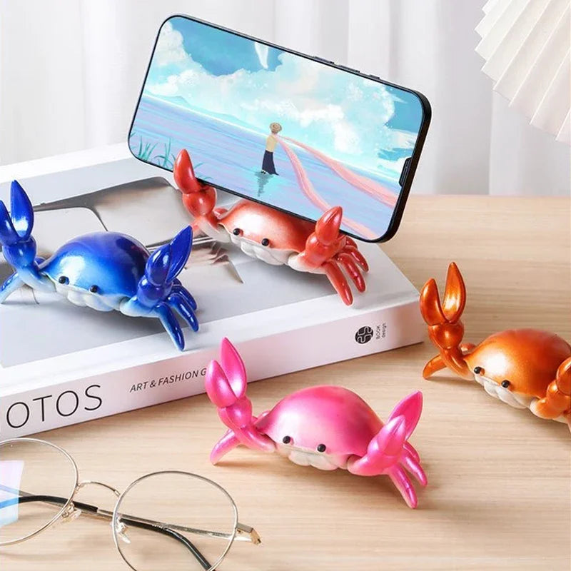 A pink crab-shaped desk organizer with a small phone holder is shown next to a white box and eyeglasses.