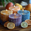 A collection of scented candles in orange, purple, and blue tones surrounded by citrus slices and fruit.