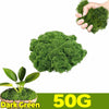 A hand is holding a clump of dark green artificial moss next to a pile of the same product.