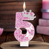 A pink glitter candle shaped like the number 5 with a crown on top, set against a wooden background.