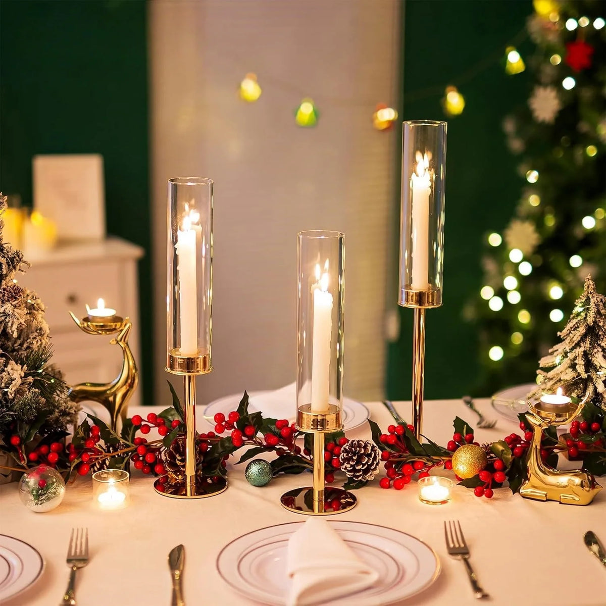 A table setting featuring gold and silver hurricane candle holders with lit candles and red foliage against a blurred Christm