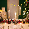 A table setting featuring gold and silver hurricane candle holders with lit candles and festive greenery.