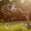 A hand is blowing a stream of liquid across many iridescent soap bubbles in a sunny outdoor setting.