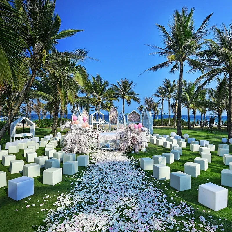 A green lawn decorated with white and pink artificial rose petals leading to a wedding archway surrounded by palm trees.