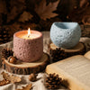 A glowing candle in a pink textured silicone mold sits on a wooden slab surrounded by pinecones and leaves.
