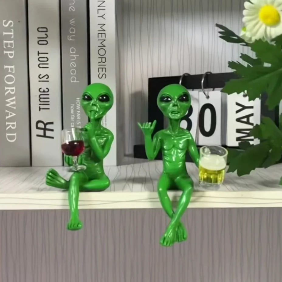 A green resin alien statue with a beer mug sits on a white shelf next to books and a flower.