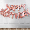 A pink foil letter birthday balloon banner spelling out HAPPY BIRTHDAY hanging on a white door.