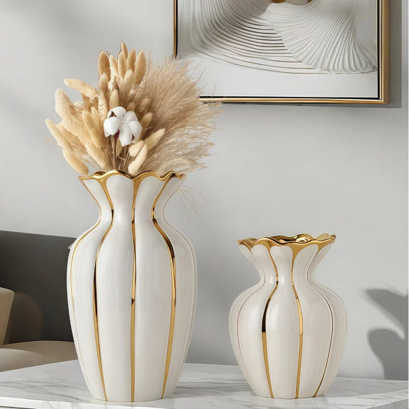 A white ceramic flower vase with gold accents sits on a marble surface, decorated with dried beige grasses.