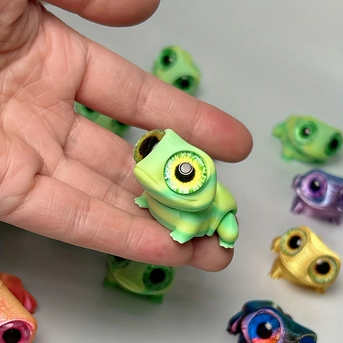 A hand holds several 3D printed frog car ornaments with color-shifting eyes in various shades of green and purple.