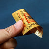 A hand holds a gold foil sticker with Chinese characters for lucky wealth and prosperity against a dark blue background.