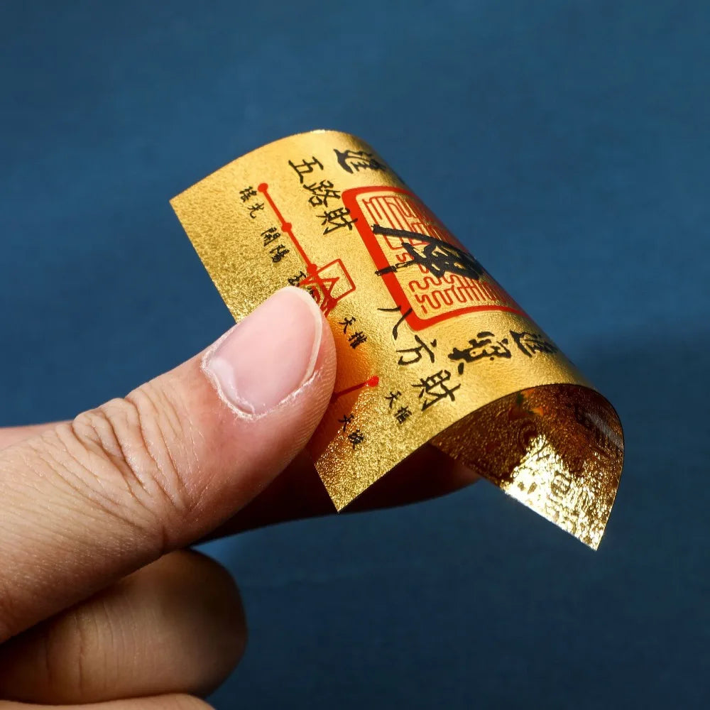 A hand holds a gold foil sticker with Chinese characters for lucky wealth and prosperity against a dark blue background.