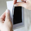 Hands are placing a black photo frame containing a handprint onto a white wall surface.