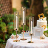 A display of three hurricane candle holders with white candles on a pink tablecloth in an outdoor setting.