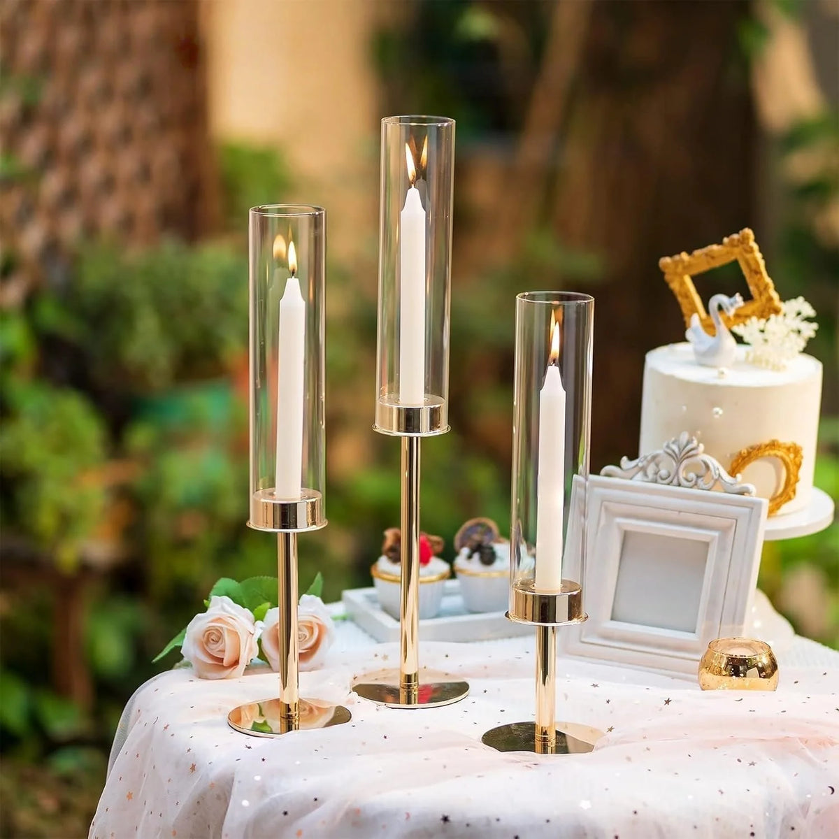 A display of three hurricane candle holders with white candles on a pink tablecloth in an outdoor setting.
