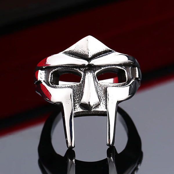 A silver stainless steel mask ring featuring a stylized face design, displayed on a reflective surface.