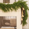 A long garland of artificial Norfolk pine greenery with gold bells hangs over a fireplace mantel.