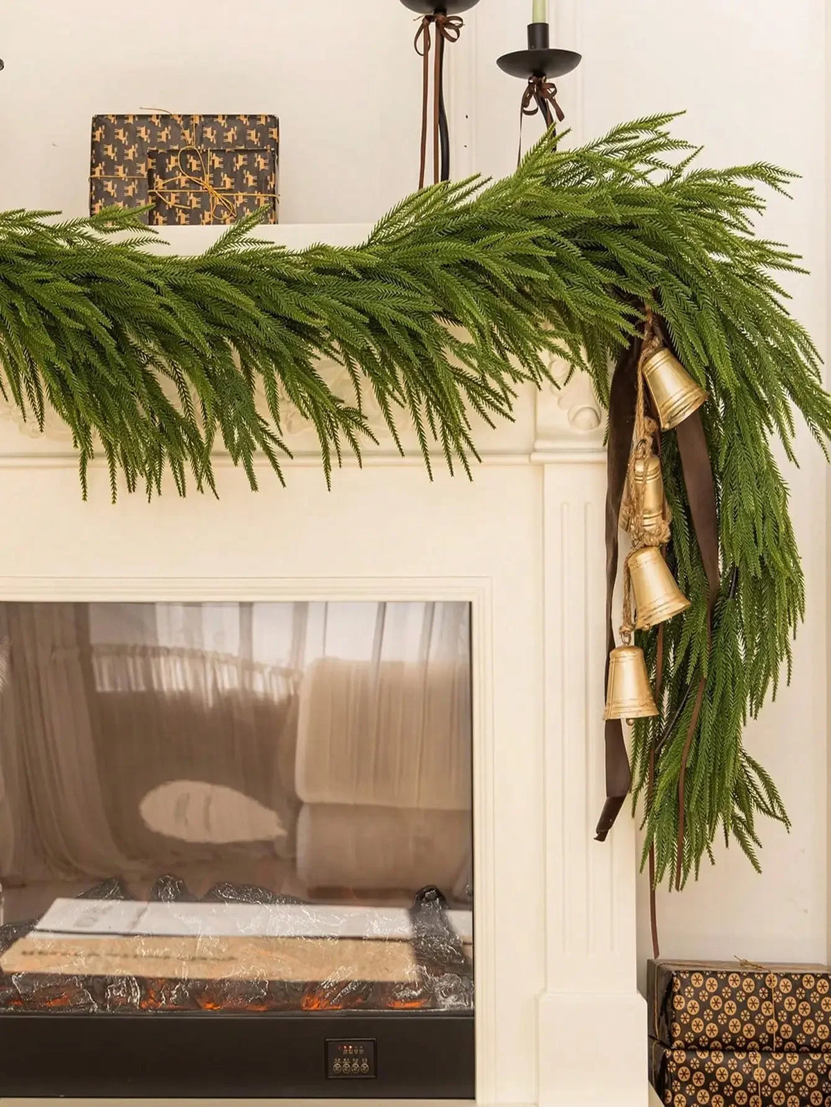 A long garland of artificial Norfolk pine greenery with gold bells hangs over a fireplace mantel.