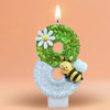 A birthday candle shaped like the number 8, decorated with a white daisy and a bee on a blue frosted base.