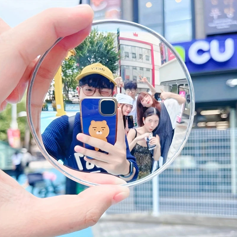 A hand holds a round convex mirror showing a person wearing a yellow cap and glasses in an outdoor setting.