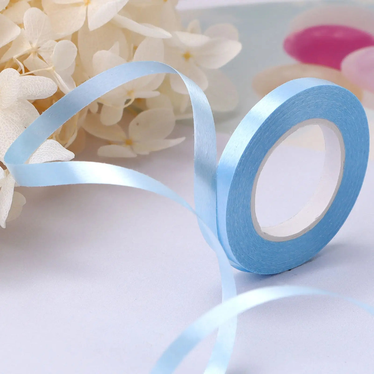 A roll of light blue satin balloon ribbon unwinding on a white surface next to white flowers and pink balloons.