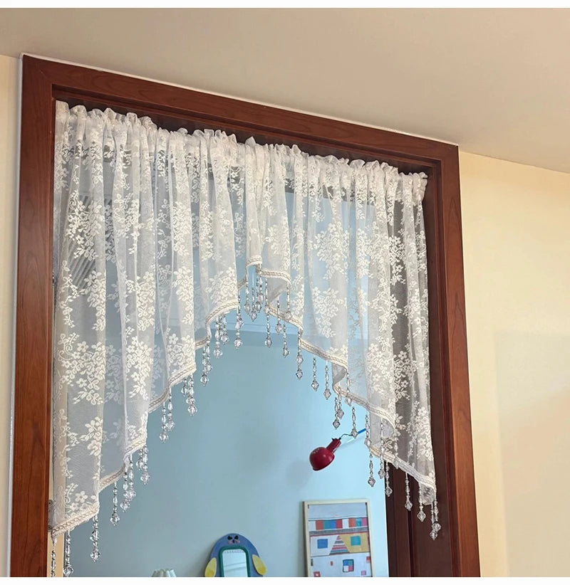 A sheer white beaded lace valance curtain hanging in a wooden frame, displayed in a room setting.