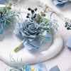 A close-up of a dusty blue silk rose boutonniere set featuring various floral and greenery elements on a white plate.