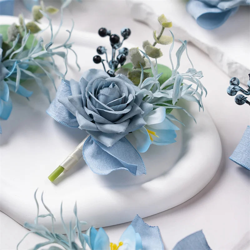 A close-up of a dusty blue silk rose boutonniere set featuring various floral and greenery elements on a white plate.