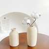 A minimalist ceramic vase in a light beige tone holds a cluster of fluffy white cotton balls against a white background.