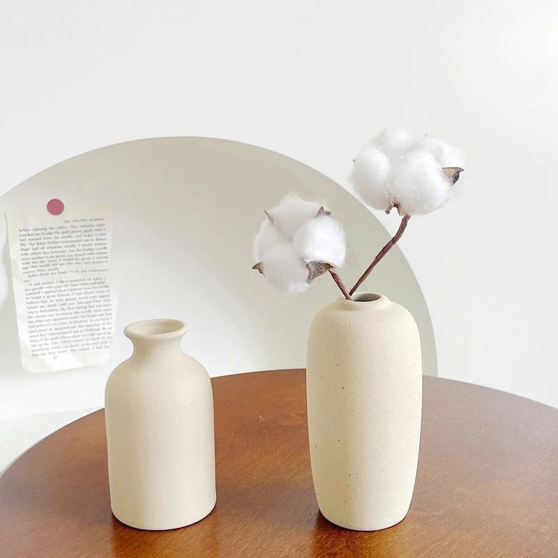 A minimalist ceramic vase in a light beige tone holds a cluster of fluffy white cotton balls against a white background.