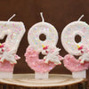 Tres velas blancas con forma de los números 7, 8 y 9 decoradas con purpurina rosa y pequeñas figuras de unicornios blancos.