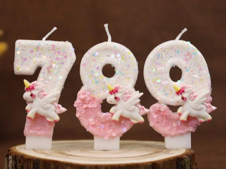 Tres velas blancas con forma de los números 7, 8 y 9 decoradas con purpurina rosa y pequeñas figuras de unicornios blancos.