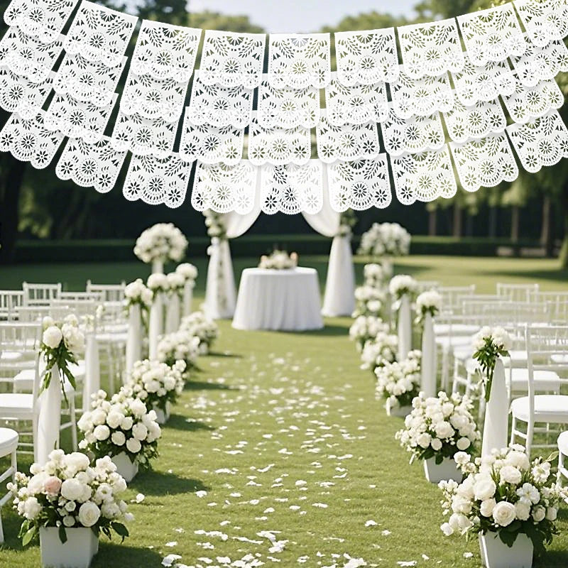 Weißer handgeschnittener Papel-Picado-Banner hängt über einer grünen Rasenfläche, dekoriert mit weißen Blumenarrangements und