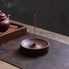 A dark brown solid wood incense stick burner with a thin metal stick emitting smoke on a dark surface.