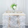 Un camino de mesa de encaje blanco cubre una mesa con un jarrón de flores moradas y blancas y copas de champán.