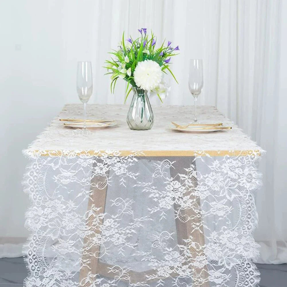 Un camino de mesa de encaje blanco cubre una mesa con un jarrón de flores moradas y blancas y copas de champán.