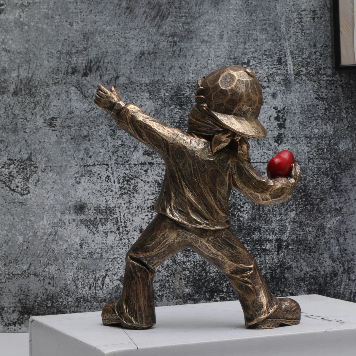 A bronze-colored resin sculpture of a figure throwing a red ball against a textured gray background.
