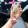 A hand holds a peach heart crystal keychain charm with a silver clasp and a light beige leather strap.