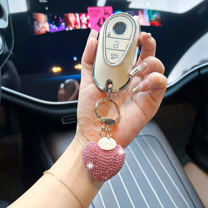 A hand holds a peach heart crystal keychain charm with a silver clasp and a light beige leather strap.