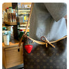 A brown leather handbag with a gold strap and a red strawberry charm is shown hanging on a person's shoulder.