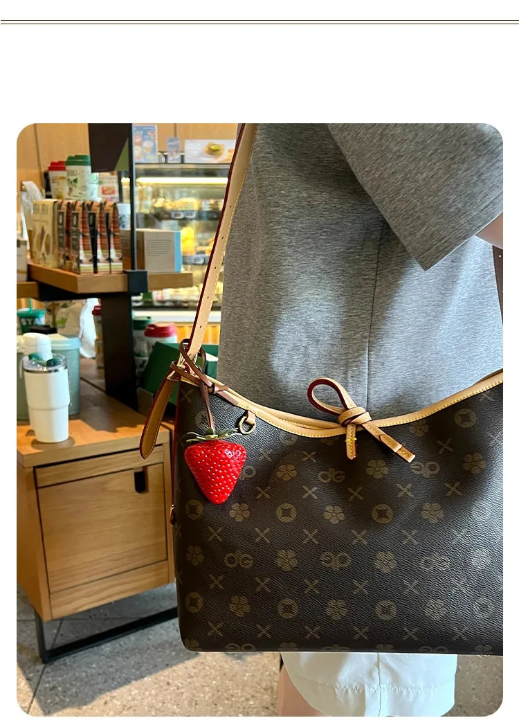 A brown leather handbag with a gold strap and a red strawberry charm is shown hanging on a person's shoulder.