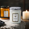 A white jar of luxurious scented candle with a patterned lid sits on a textured fabric next to a lit candle.
