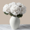 A white ceramic vase holds a large, fluffy white hydrangeas flower arrangement.