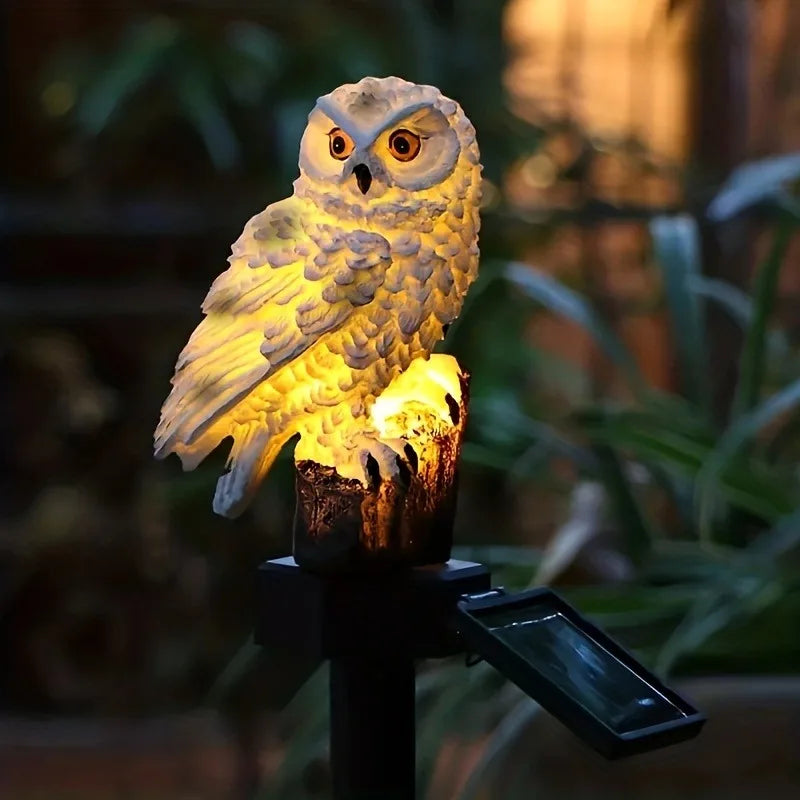 A realistic owl sculpture illuminated by a solar light, positioned on a dark stake in a garden setting.