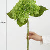 A green hydrangea silk flower branch with a stem and leaves is shown against a white background.