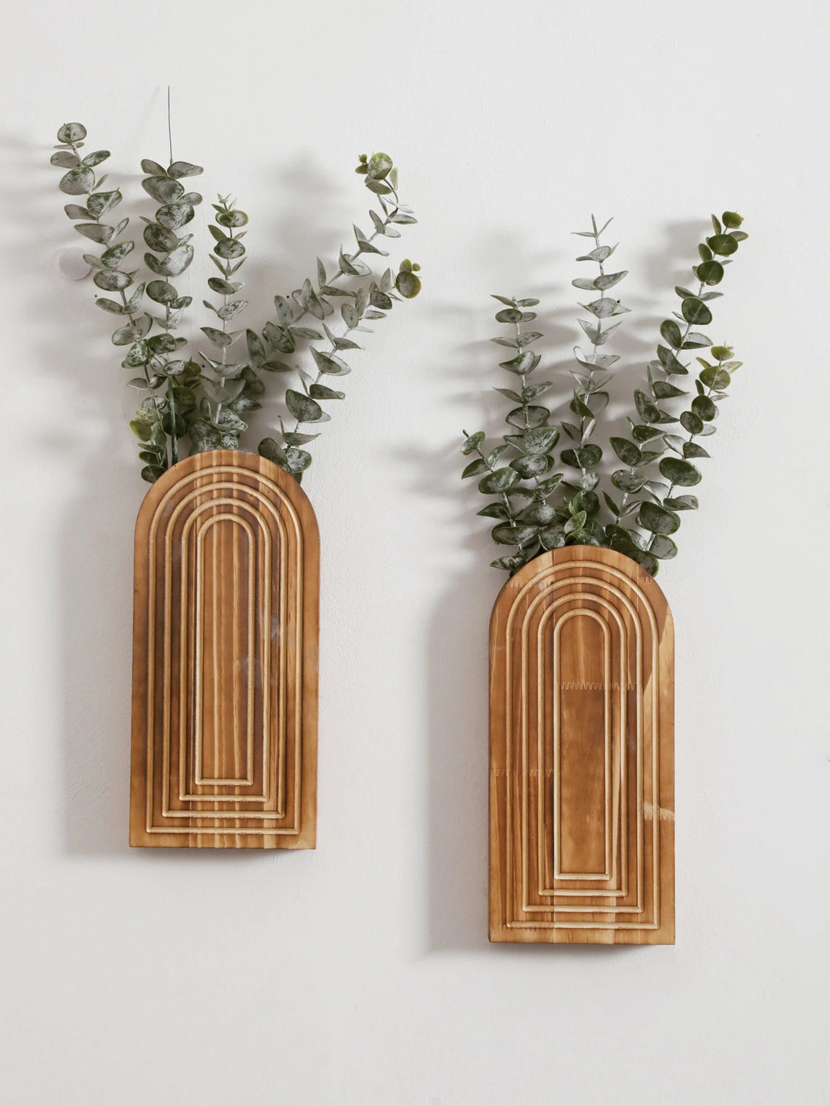 Un par de jarrones de madera arqueados con ramas de eucalipto verde montados en una pared blanca.