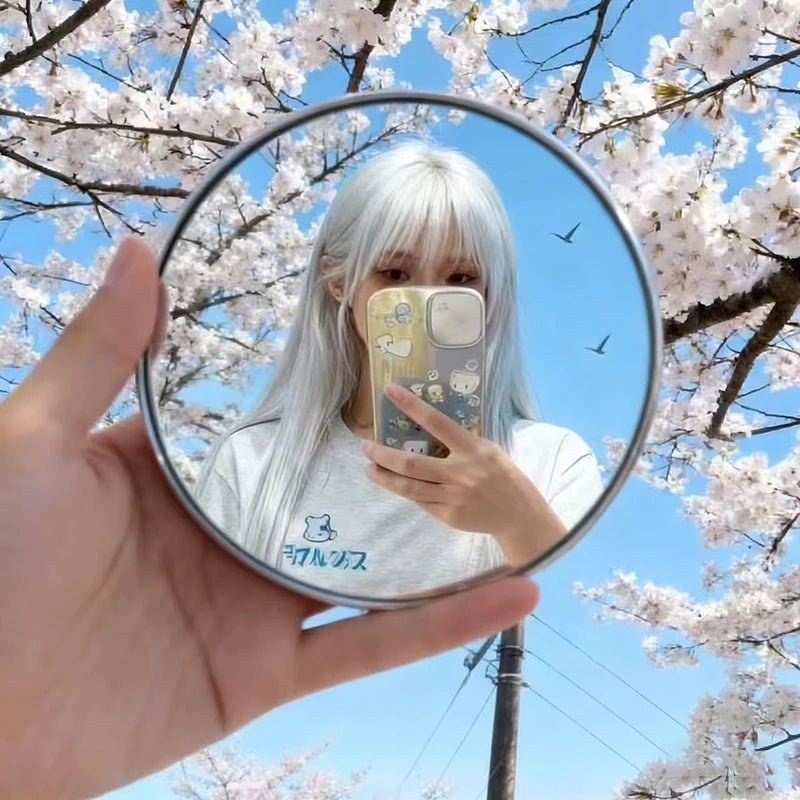 A hand holds a round convex mirror showing a person with long white hair and a light gray shirt against a background of pink 
