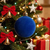 A close-up of a deep blue velvet flocked Christmas ornament hanging on a green fir tree with red bows and gold accents.