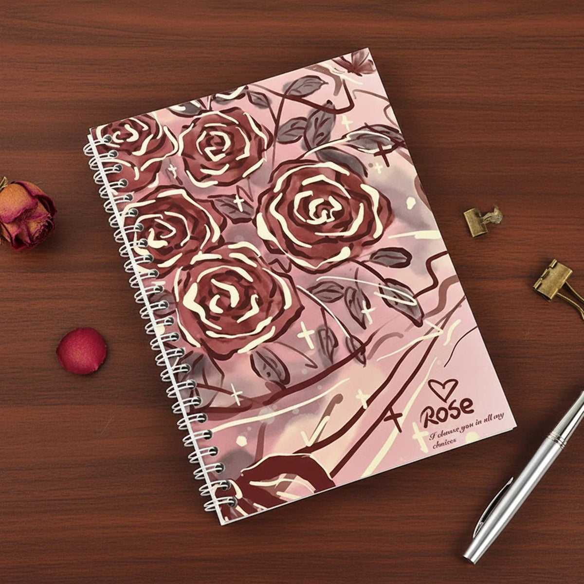 A spiral-bound notebook featuring a floral pattern of roses and leaves, lying on a wooden surface with a pen and small metal 